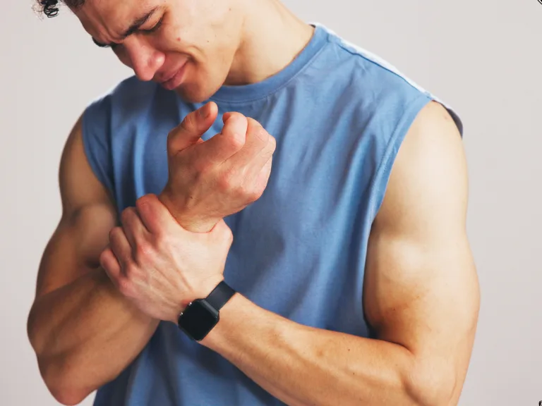 Man in a blue shirt holding his wrist in discomfort against a plain background.